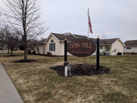View of the exterior of Ives Hill Retirement Community building with a sign in the foreground and an American flag on a flagpole behind it. The building is a single-story structure with beige siding and several windows. There are leafless trees and a grassy area surrounding the building.