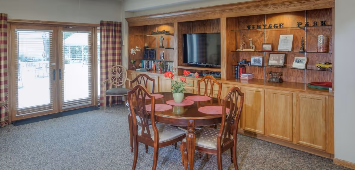 A cozy common area with a round wooden table and four chairs, each with a cushioned seat. The table has a green potted plant with red flowers and four pink placemats. Behind the table is a built-in wooden shelving unit with a flat-screen TV, decorative items, framed photos, and books. To the left, there are large glass doors with wooden frames and plaid curtains, leading to an outdoor patio area with tables and chairs.