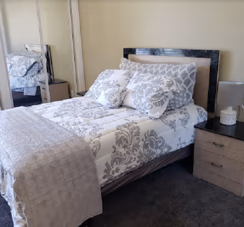 A neatly made bed with a patterned white and gray comforter and matching pillows in a bedroom. There is a bedside table with a lamp and a mirrored wardrobe reflecting part of the bed.
