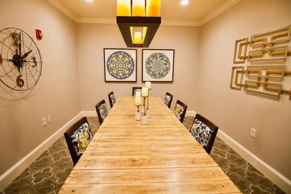 A dining room with a long wooden table surrounded by eight chairs with floral patterned cushions. The table has three candle holders with candles. On the wall at the far end are two framed circular patterned artworks. A large decorative clock is mounted on the left wall, and a geometric wall art piece is on the right wall. The room is warmly lit by a rectangular ceiling light fixture.