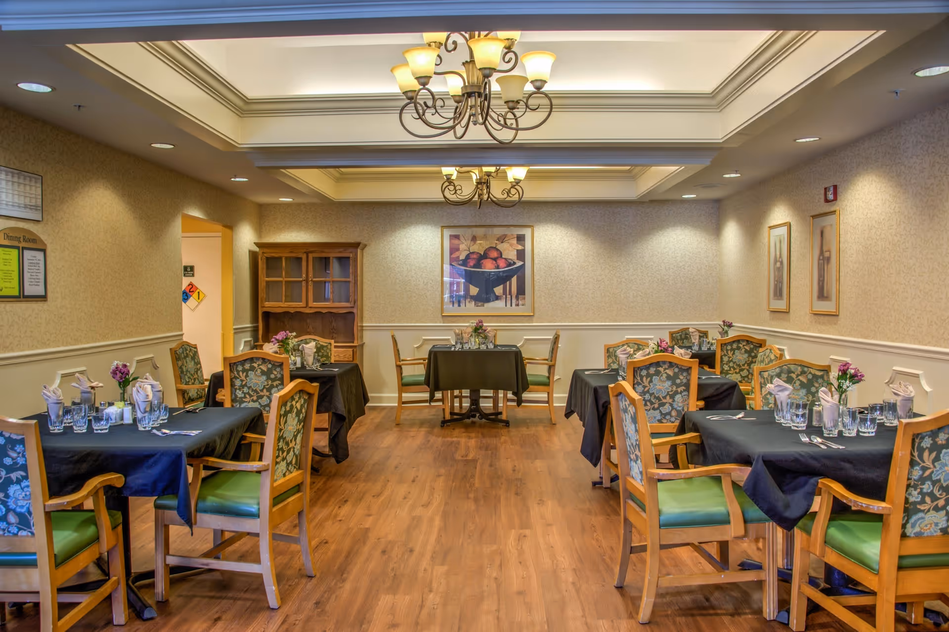 A dining room with several tables covered in dark tablecloths, each set with glasses, napkins, and silverware. The chairs have wooden frames with green cushioned seats and floral-patterned backs. The room has wooden flooring, beige walls with white wainscoting, and decorative ceiling lighting fixtures. There are framed pictures on the walls and a wooden cabinet in the back corner.