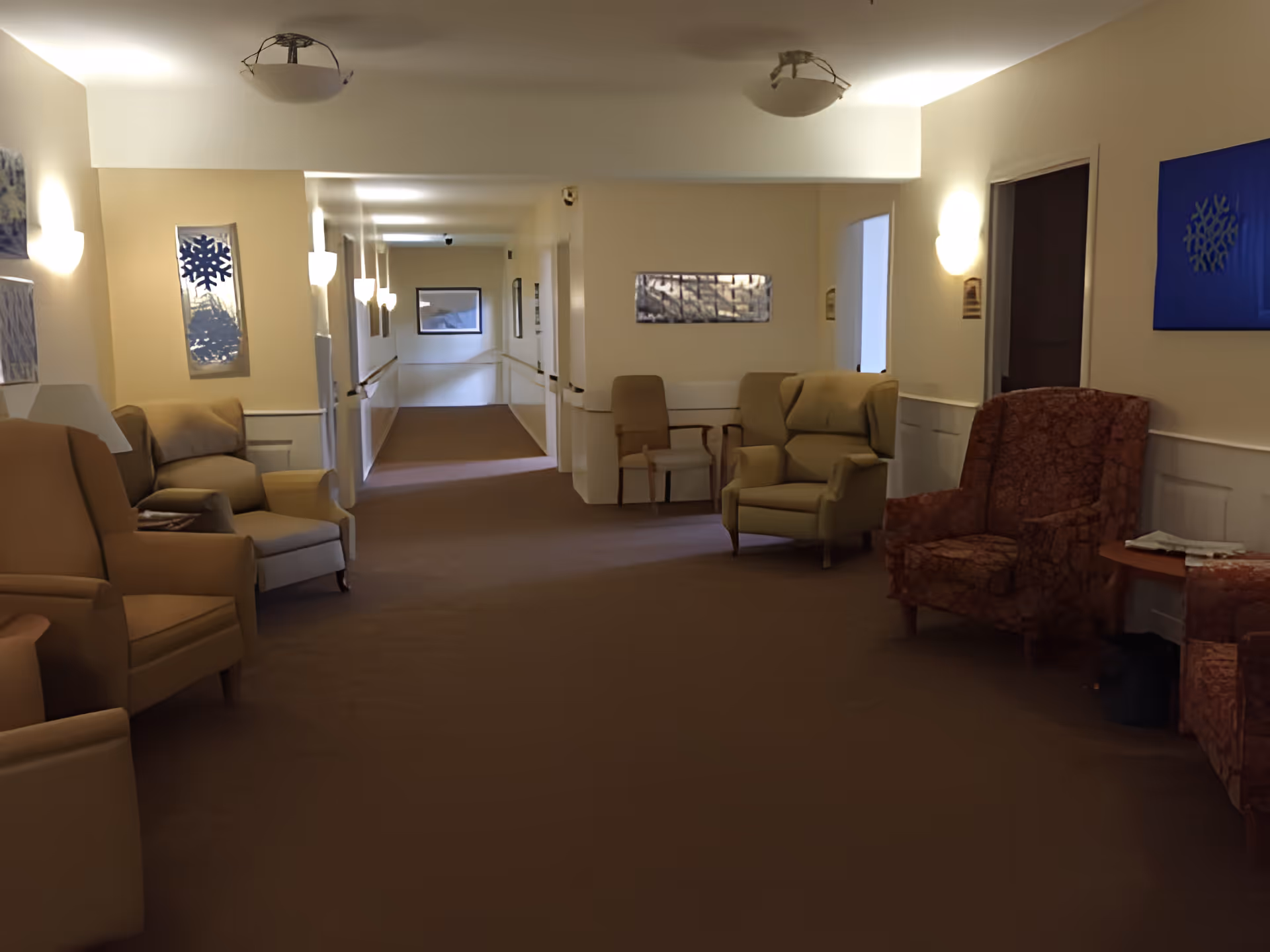 Carpeted lounge corridor with multiple armchairs and wall-mounted lights in a senior living facility common area.
