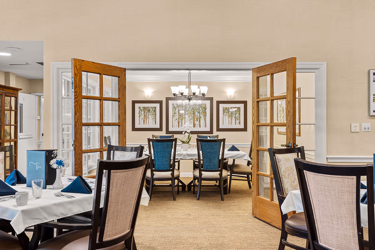 Interior view of a dining room in a senior living facility with tables set with white tablecloths, blue napkins, and glassware. Wooden chairs with cushioned seats surround the tables. French doors with glass panels open to another dining area with a chandelier and framed artwork on the walls.