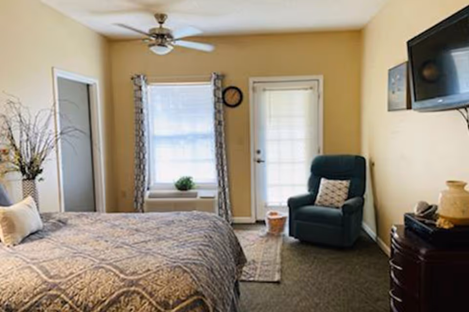 A bedroom with a large bed covered in a patterned bedspread, a ceiling fan above, a window with curtains, a door with a window, a green armchair with a pillow, a small rug, and a wall-mounted TV above a dresser with decorative items.