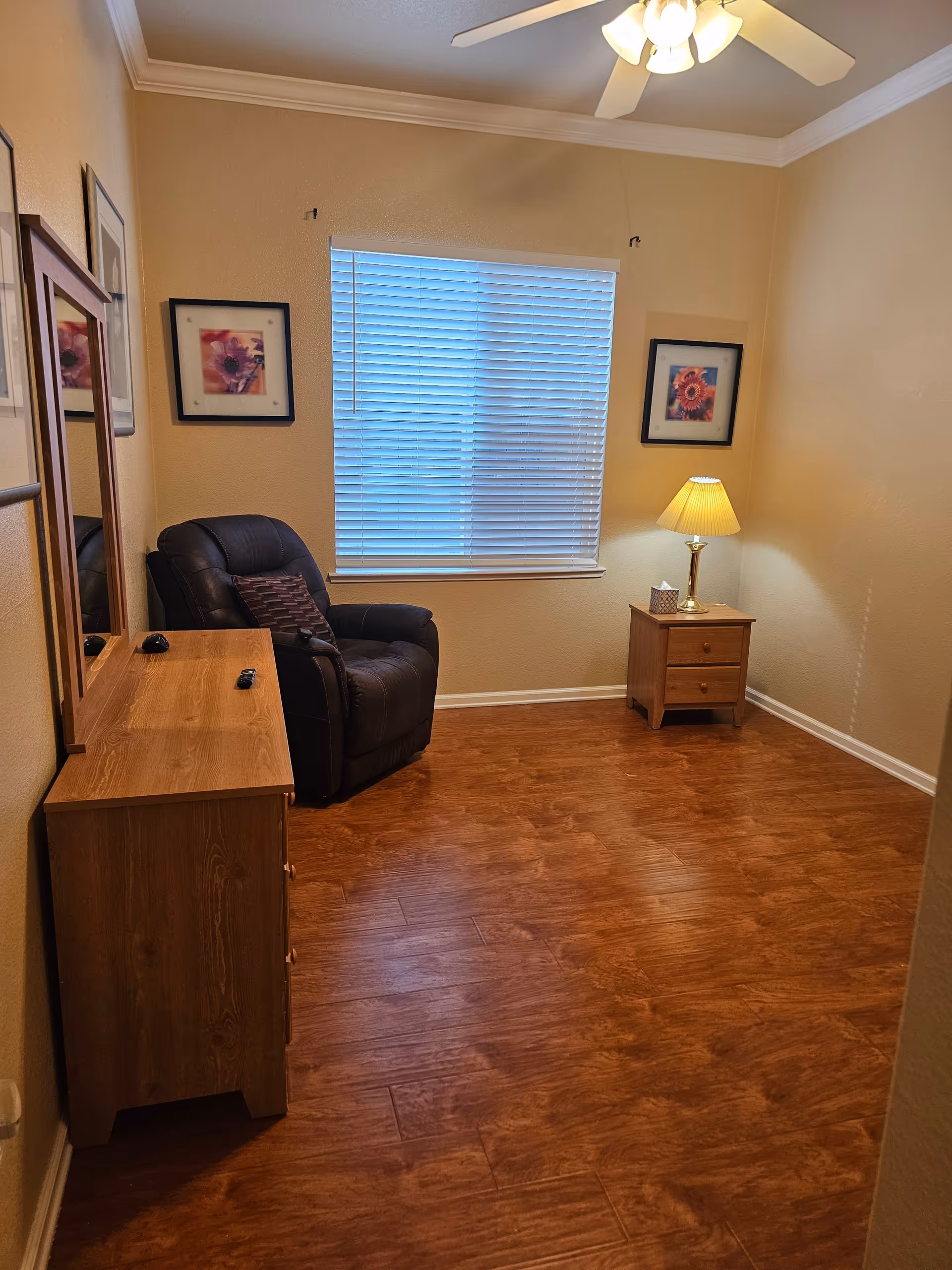 Small sitting room with a recliner, wooden dresser and nightstand with a lamp in front of a window with closed blinds.