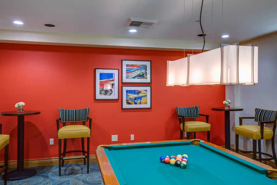 Indoor recreation room with a pool table, hanging pendant lights, red accent wall, and seating with small tables.