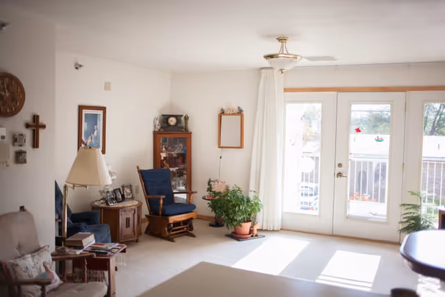 A bright living room with white walls and carpeted floor featuring a wooden rocking chair with blue cushions, a small wooden side table with framed photos, a floor lamp, a wooden cabinet with decorative items, a potted plant near glass doors leading to a balcony, and a wall clock and cross hanging on the wall.