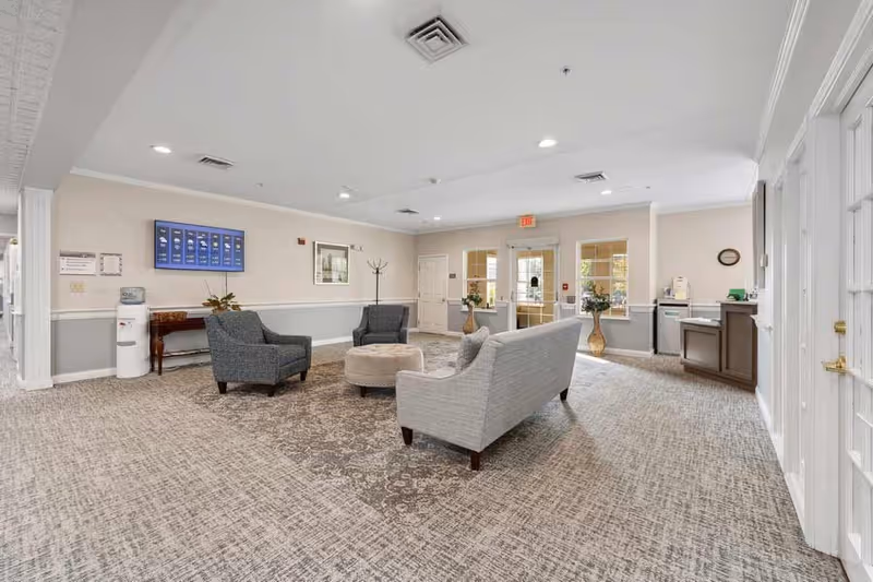 A spacious and well-lit common area in a senior living facility with light-colored walls and carpeted floor. The room features a gray sofa, two gray armchairs, and a round beige ottoman arranged in the center. There is a water cooler and a small wooden table against one wall, a coat rack, and a wall-mounted television. Large windows and glass doors allow natural light to enter the room. Decorative plants and a clock are also visible.