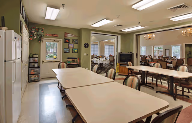 Interior view of a dining area in a senior living facility with several tables and chairs arranged for group seating. The room has green walls, a white refrigerator, and a door with a window. There are framed pictures and decorative items on the walls, and through an open doorway, another dining room with chandeliers and more tables and chairs is visible.