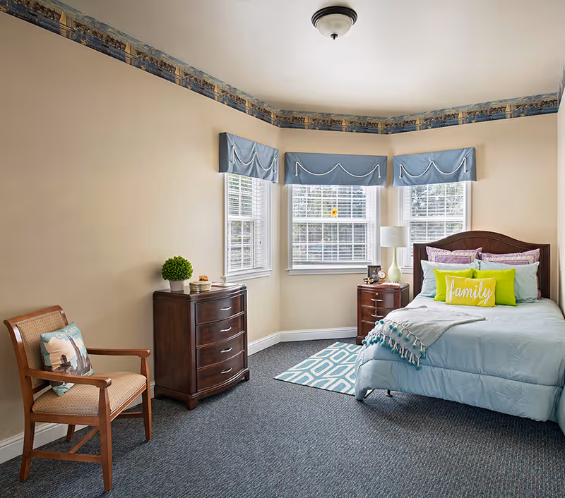 Comfortable bedroom with a blue bed, wooden dresser and nightstand, a chair, and three windows with blue valances.