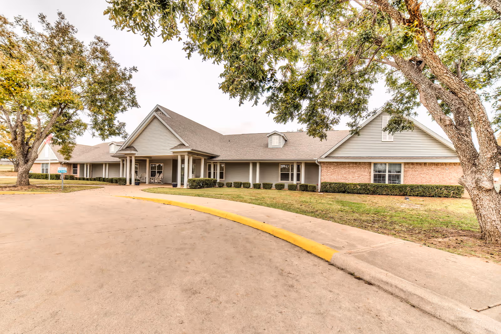Exterior view of a single-story assisted living facility building with a covered entrance, surrounded by trees and a curved driveway with a yellow curb.