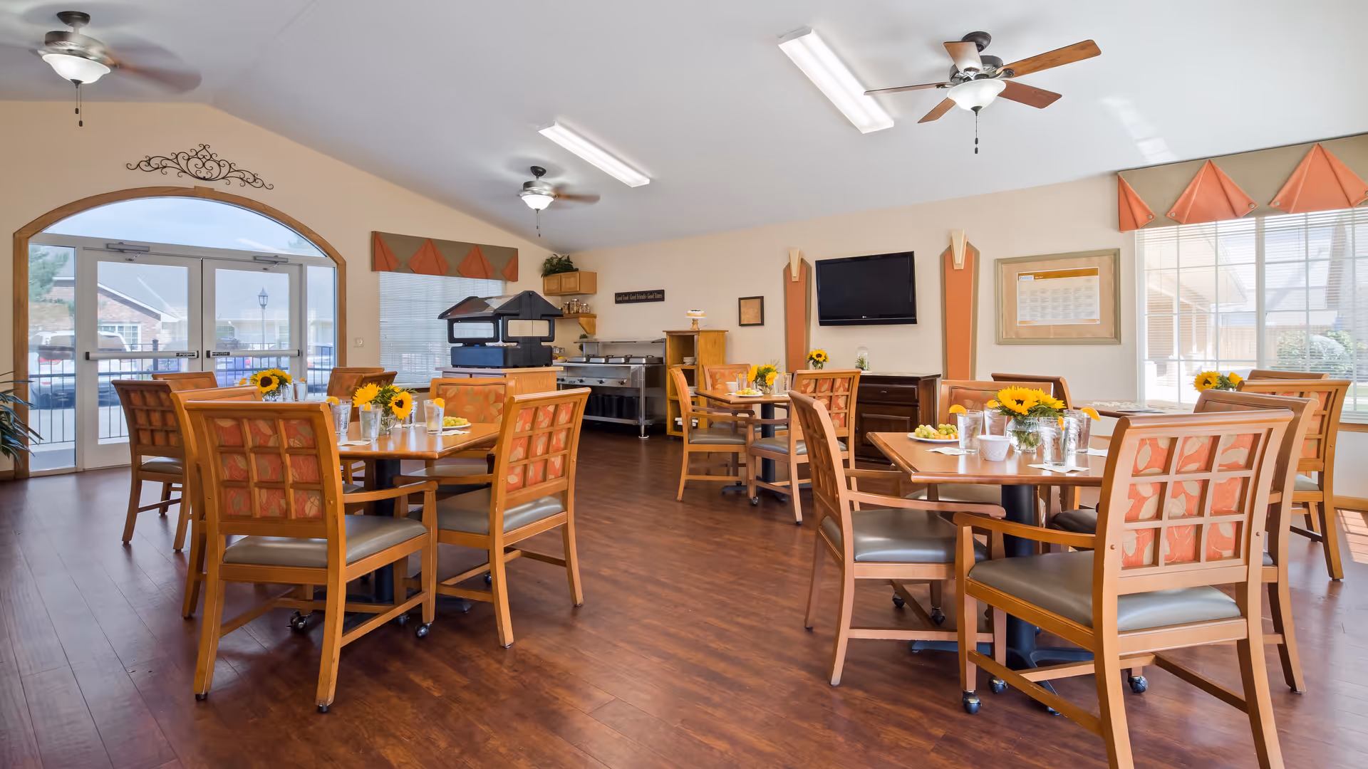 A bright dining room with wooden tables and chairs, each table decorated with a vase of sunflowers and glasses of water. The room has large windows with orange valances, ceiling fans, and a TV mounted on the wall. There is a buffet area with food warmers and a serving station in the background.