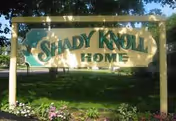 Wooden-framed Shady Knoll Home sign with teal background, set among greenery.