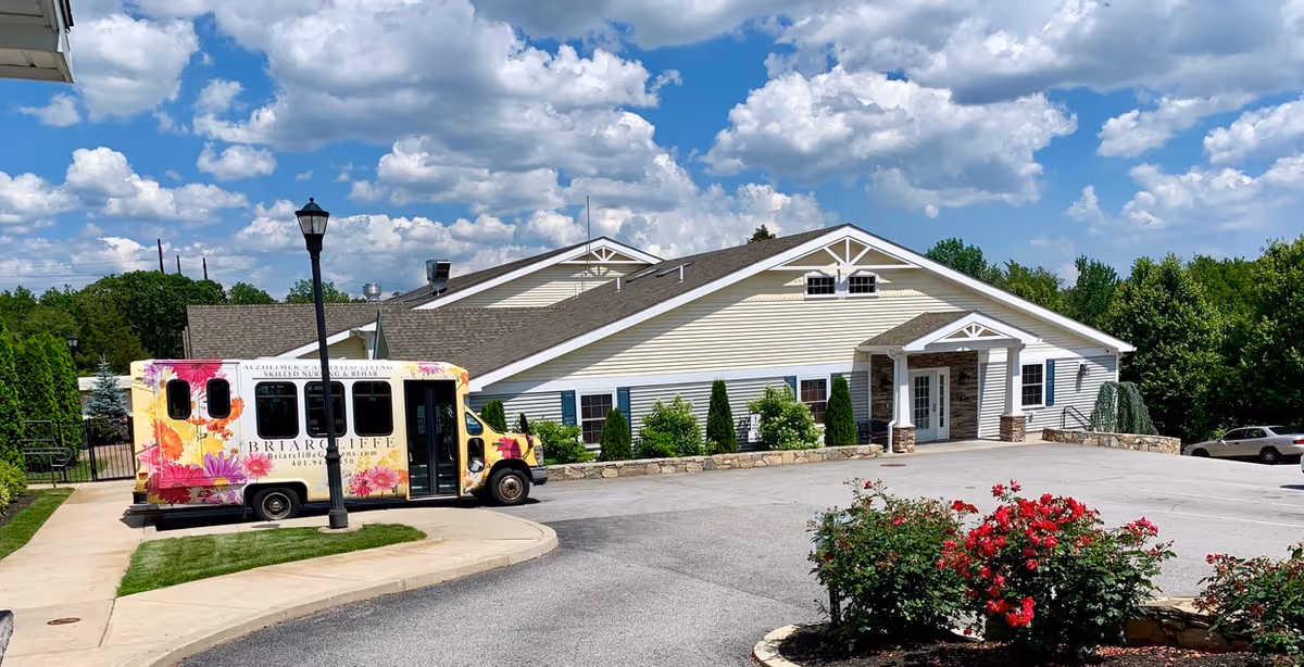 Exterior front of Briarcliffe Gardens memory care building with a floral‑branded shuttle parked at the entrance and landscaped rose bushes under a blue sky.