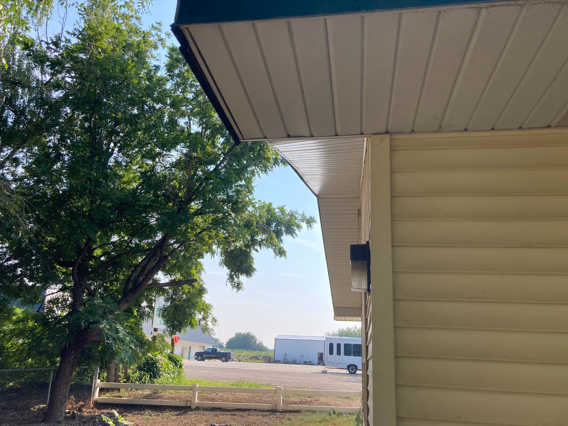 View of the side of a building with beige siding and an overhanging roof. To the left, there is a large green tree and a small white fence. In the background, there is a parking area with a black pickup truck and a white shuttle bus, along with a white building under a clear blue sky.