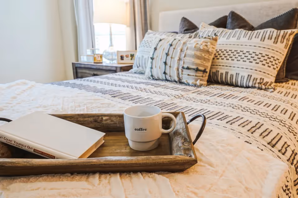 A cozy bedroom scene with a neatly made bed featuring patterned pillows and a textured blanket. On the bed is a wooden tray holding a white coffee cup labeled 'coffee' and a closed book. In the background, a nightstand with a lamp and framed photos is visible.