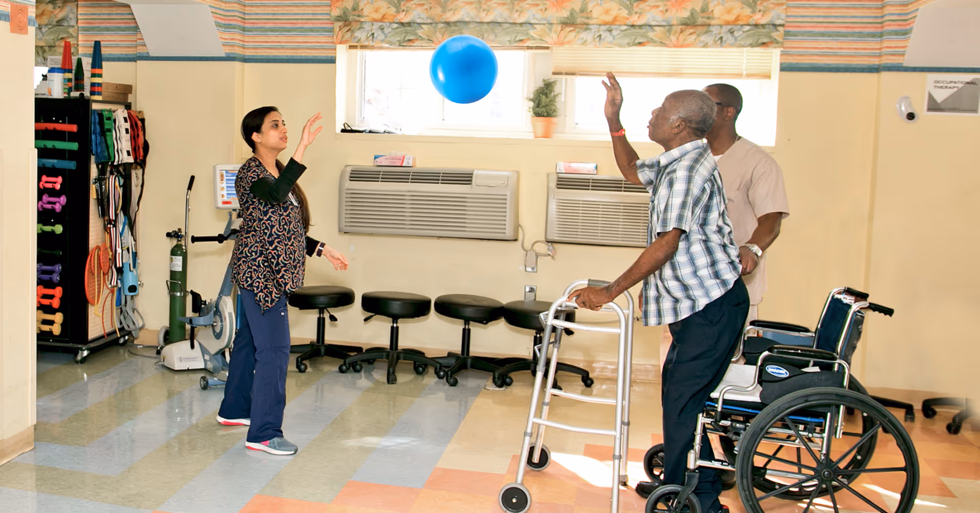 An elderly man using a walker is playing with a blue balloon with the assistance of two caregivers in a therapy room equipped with exercise equipment and stools.