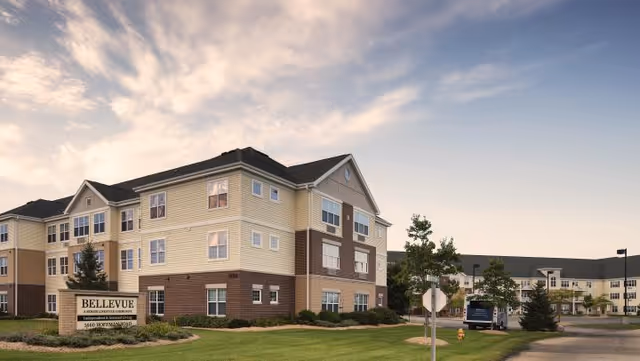 Exterior front view of a three-story Bellevue senior living building with a lawn and entrance sign under a cloudy sky.