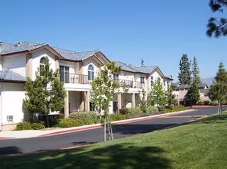 Exterior view of a two-story residential building with balconies, surrounded by trees and greenery, with a paved driveway and well-maintained lawn in the foreground under a clear blue sky.
