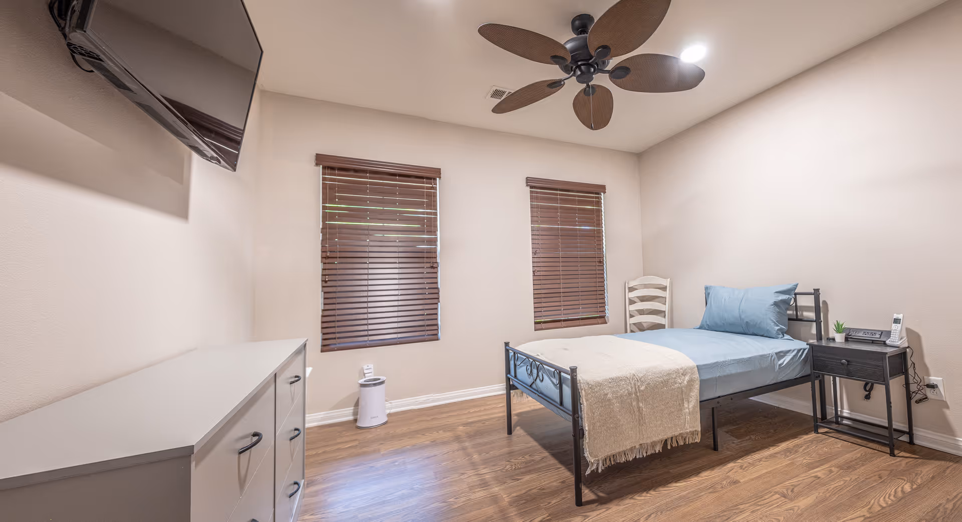 A simple furnished bedroom with a single metal bed, nightstand, dresser, wall-mounted TV, ceiling fan and two windows with blinds.