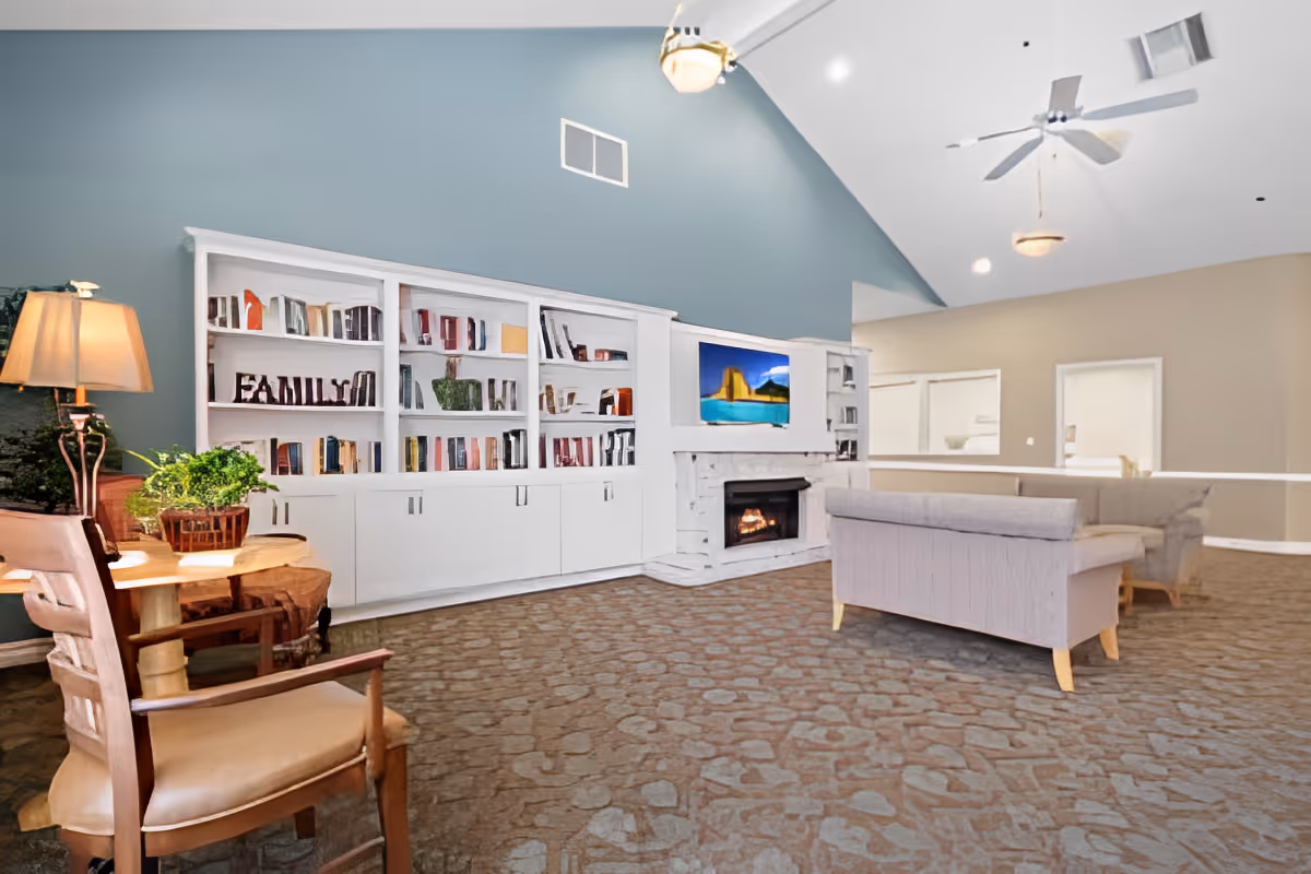 A spacious living room with a high vaulted ceiling painted blue. The room features a white built-in bookshelf filled with books and decorative items, including a sign that says 'FAMILY'. There is a fireplace with a TV mounted above it, two gray sofas facing the fireplace, a wooden chair, a round wooden table with a lamp and a potted plant. The floor has a stone-patterned carpet.