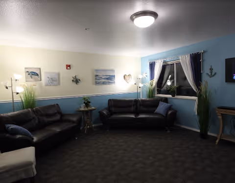 A cozy lounge with two dark leather sofas, side tables, floor lamps, wall art and a curtained window.