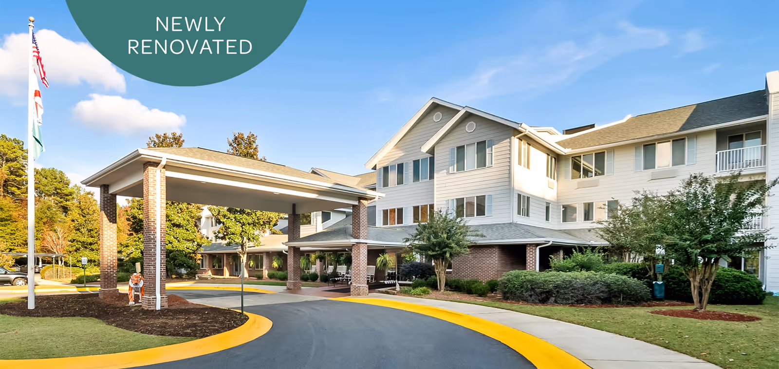 Front exterior of a three-story senior living building with a covered drop-off porte-cochere, flags, and landscaped grounds under a 'Newly Renovated' banner.