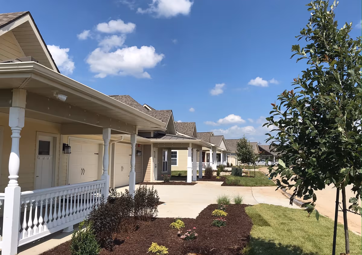 Exterior view of a row of single-story residential buildings with covered porches, white railings, and garages. There are small landscaped areas with bushes and young trees along a paved walkway under a blue sky with scattered clouds.