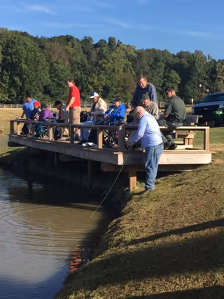 A group of elderly people and caregivers fishing off a wooden dock by a pond on a sunny day, with trees and a clear blue sky in the background.