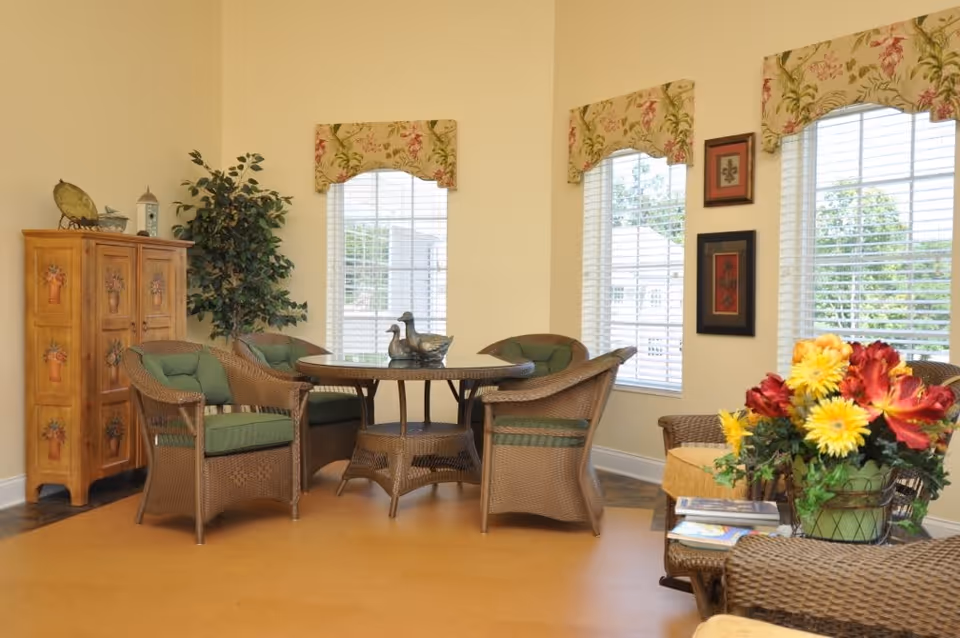 A cozy sitting area with a round wicker table surrounded by four wicker chairs with green cushions. Behind the table are three windows with floral valances, letting in natural light. To the left, there is a wooden cabinet with floral designs and a tall potted plant. On the right, a wicker armchair with a yellow cushion and a side table holding a colorful flower arrangement and some magazines are visible. The walls are painted a soft yellow.