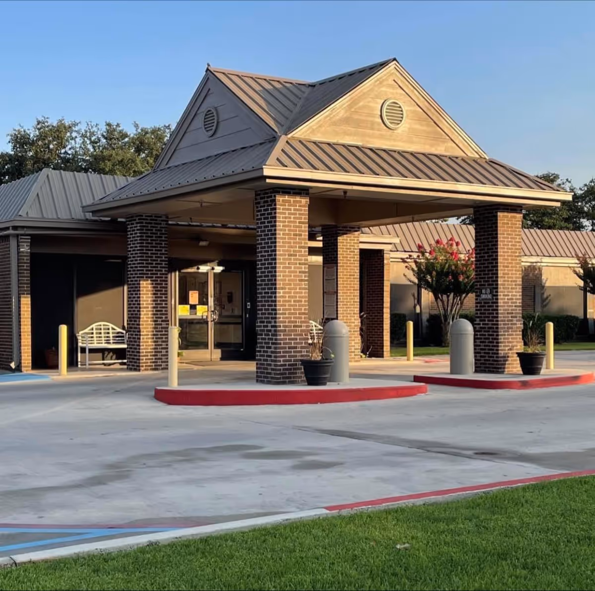Exterior view of Brownwood Nursing & Rehabilitation facility entrance with a covered drop-off area supported by brick columns, a bench to the left, potted plants, and a clear sky.