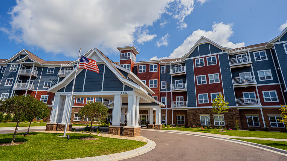 Entrance and front facade of a multi-story senior living building with a covered porte-cochere and an American flag.