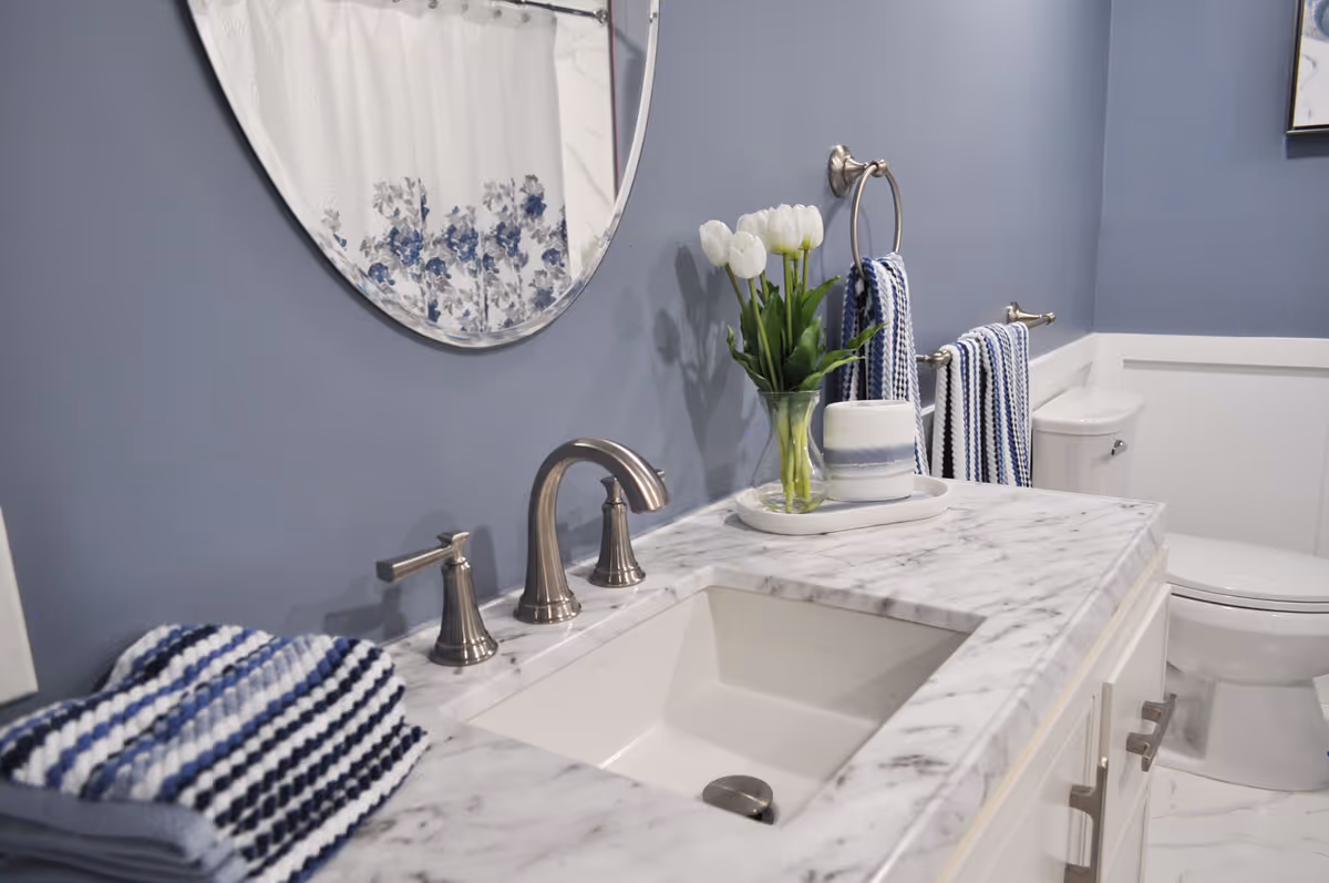A bathroom with a marble countertop sink, silver faucet, and a round mirror above. The walls are painted blue with white wainscoting. There are white tulips in a glass vase and a decorative container on a tray next to the sink. Blue and white striped towels hang on silver towel rings and bars. A white toilet is visible in the background.