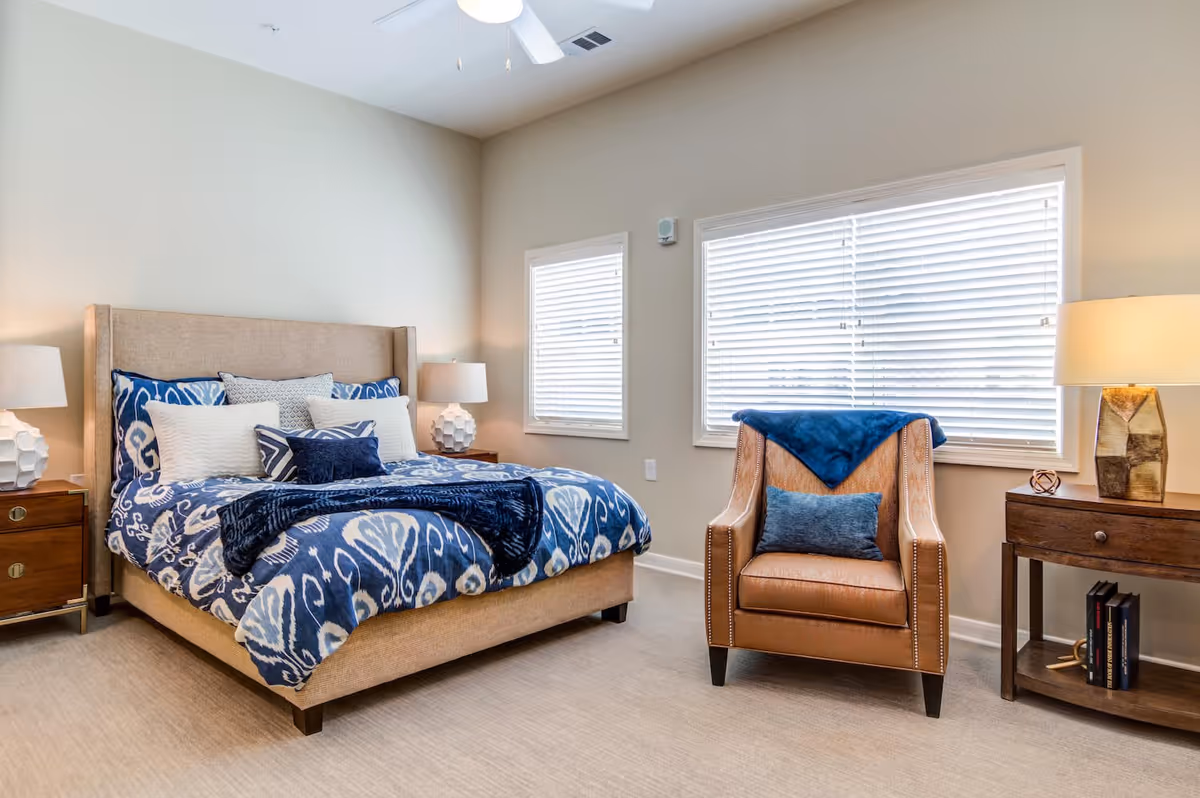 A well-lit bedroom featuring a beige upholstered bed with blue and white patterned bedding and multiple pillows. On either side of the bed are wooden nightstands with white lamps. To the right, there is a brown leather armchair with a blue throw blanket and a blue cushion. The room has two windows with white blinds and neutral-colored walls and carpet.