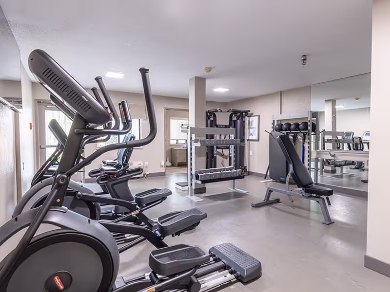 A fitness room with exercise equipment including elliptical machines, a weight bench, dumbbells on a rack, and a mirrored wall reflecting the room.