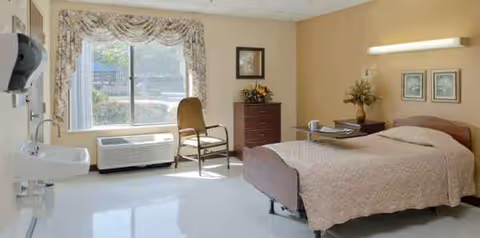 A senior living bedroom with a bed, chair, dresser, and window in view.