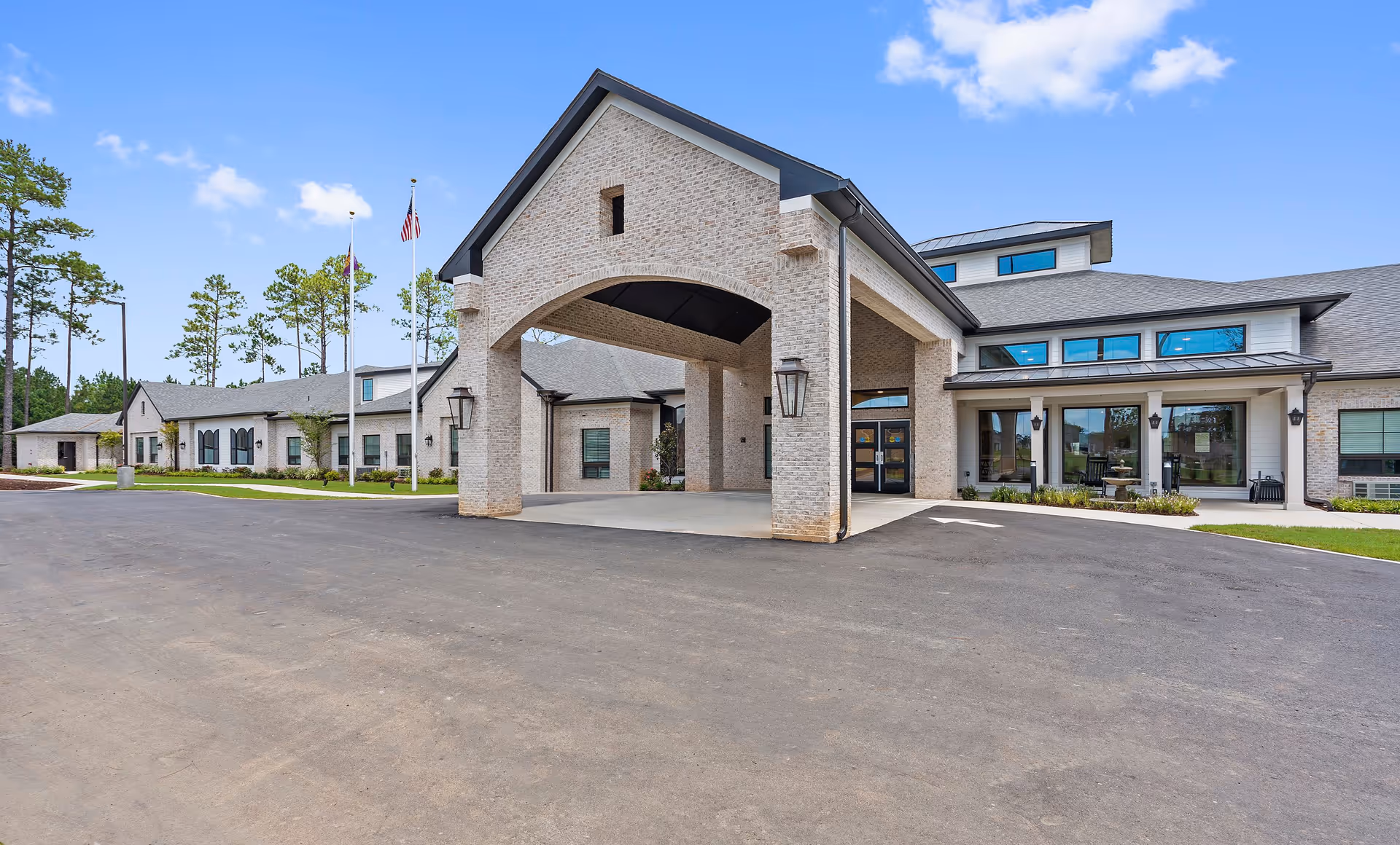 Exterior view of Sage Lake senior living facility showing a large covered entrance with brick pillars, multiple windows, and a paved driveway. There are three flagpoles with flags in front of the building and trees in the background under a blue sky with some clouds.