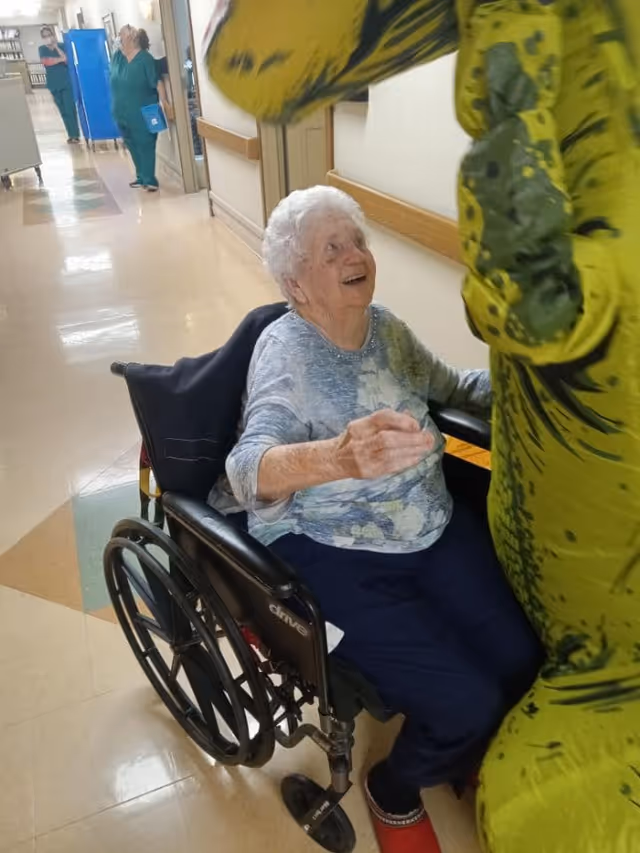 An elderly woman in a wheelchair smiling and reaching out towards a person dressed in a green dinosaur costume in a hallway of a healthcare facility. In the background, two healthcare workers in green scrubs are standing and talking.