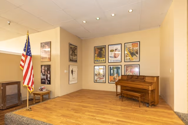 A small stage area with a wooden floor and a piano with a bench on the right side. An American flag stands on the left side of the stage next to a small table and a speaker. The back wall is decorated with framed vintage posters.