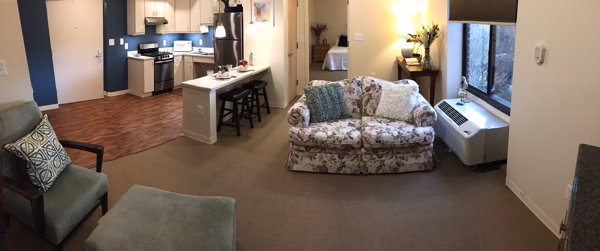 Interior view of a senior living apartment showing a living area with a floral patterned loveseat, a green armchair with a decorative pillow, and an ottoman. The room has a large window with a heating/cooling unit beneath it. In the background, there is a kitchen area with white cabinets, a stove, microwave, and refrigerator, along with a breakfast bar with two stools. A bedroom is visible through an open doorway.