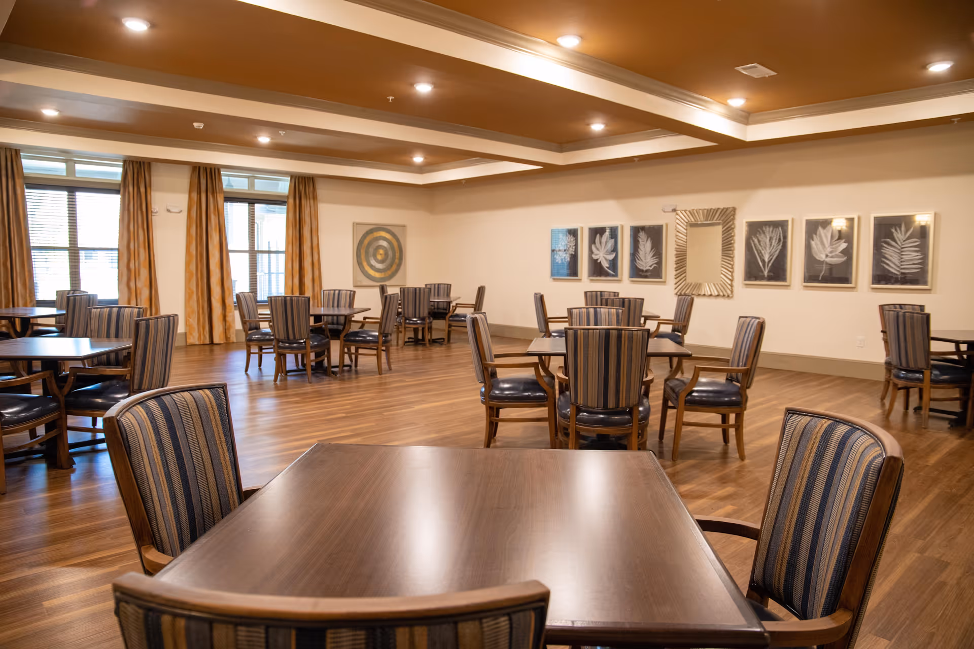 A spacious dining room with multiple wooden tables and striped cushioned chairs arranged neatly. The room features large windows with yellow curtains, wooden flooring, and framed botanical artwork on the walls. The ceiling has recessed lighting and a tray design.