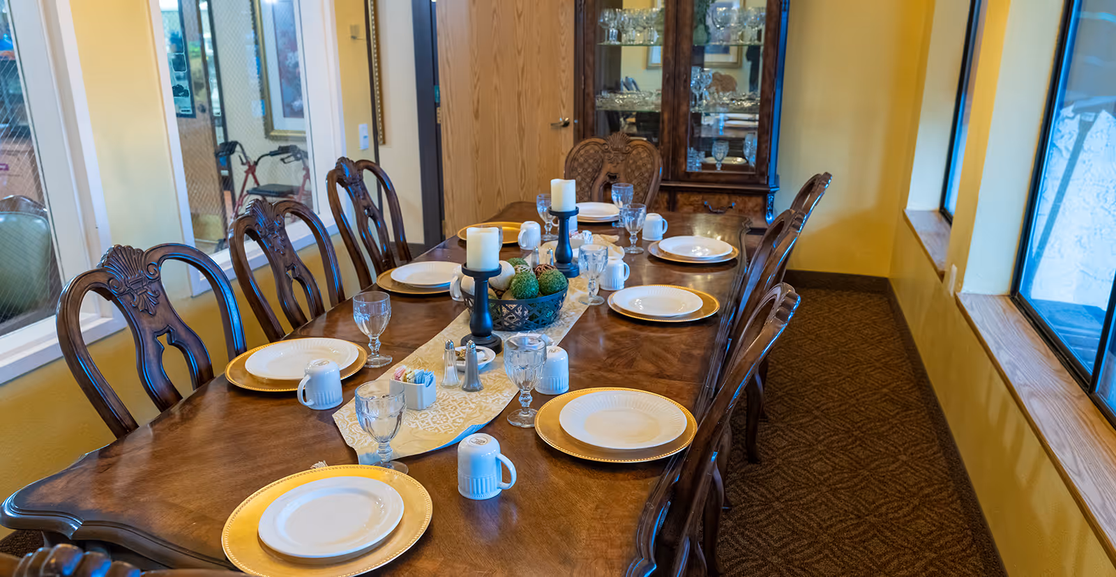 Long wooden dining table set with plates, glassware, candles and a decorative centerpiece surrounded by carved chairs in a dining room.