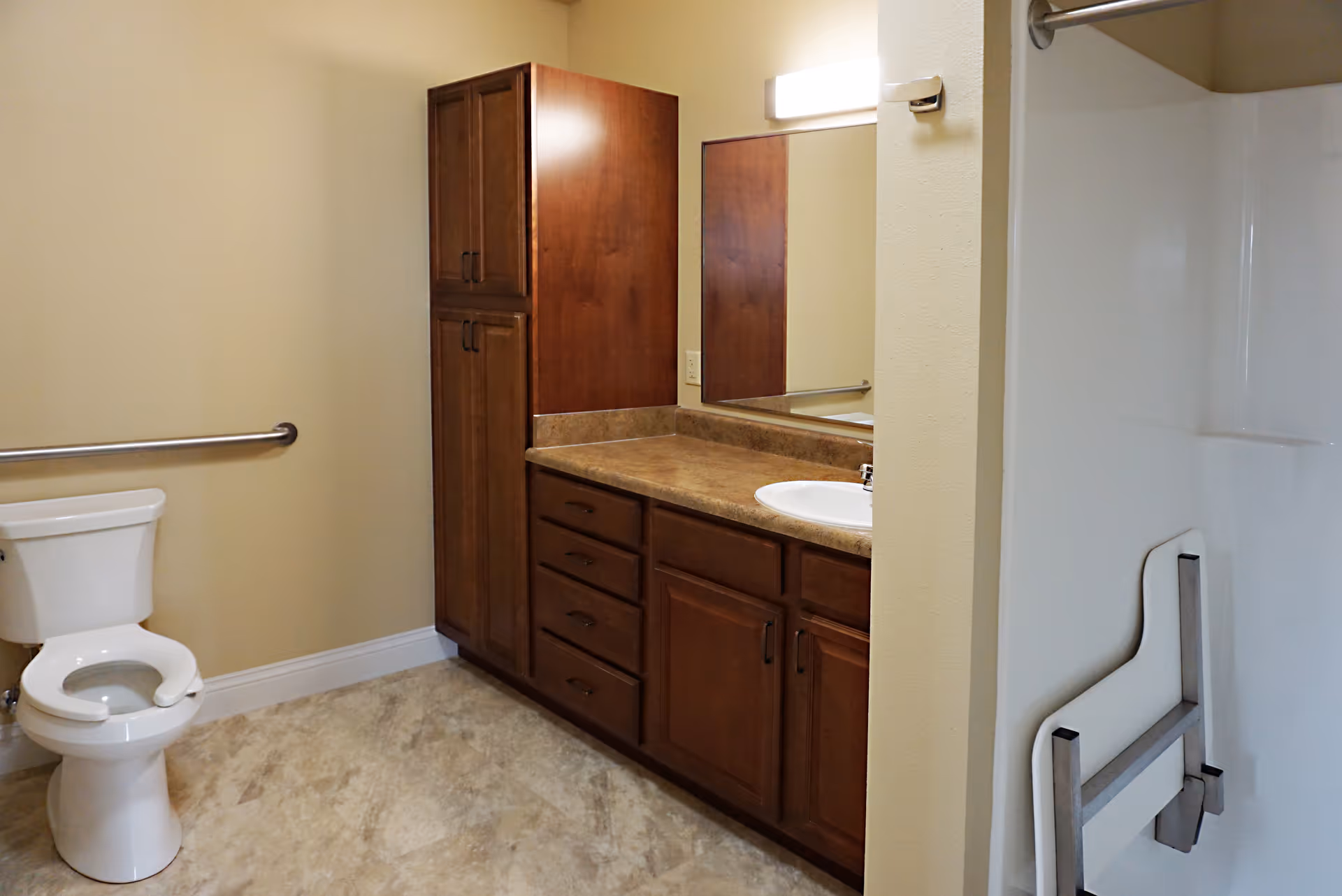 A bathroom with a white toilet, a wooden cabinet with multiple drawers and doors, a countertop with a sink, a large mirror above the sink, and a grab bar on the wall next to the toilet. The floor has a light-colored tile and the walls are painted beige.