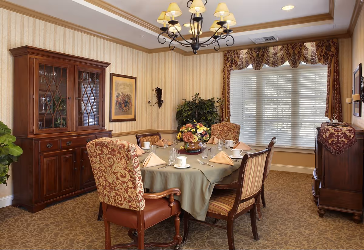 A warmly decorated dining room with a round table set for six people. The table is covered with a green tablecloth and has beige napkins, white cups, glasses, and silverware. The room features patterned upholstered chairs, a wooden china cabinet with glass doors displaying glassware, a sideboard with a decorative cloth, a large window with blinds and ornate curtains, a chandelier with multiple lampshades, framed artwork on the walls, and a potted plant in the corner.