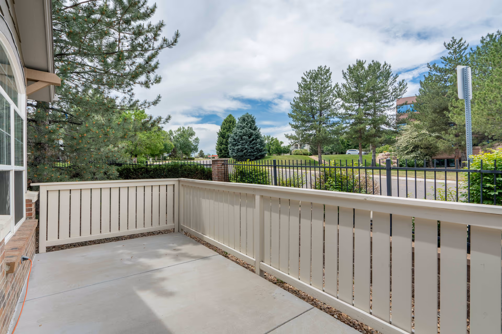 A small outdoor patio area with a concrete floor and beige wooden fencing. The patio is adjacent to a brick building with large windows. Beyond the fence, there are trees, a road, and a partly cloudy sky.