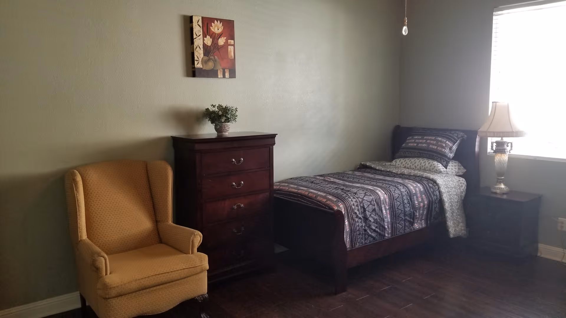 A simple bedroom with a single bed covered in patterned bedding, a wooden nightstand with a decorative lamp, a wooden chest of drawers with a small potted plant on top, a yellow upholstered armchair, and a framed floral painting on the wall.