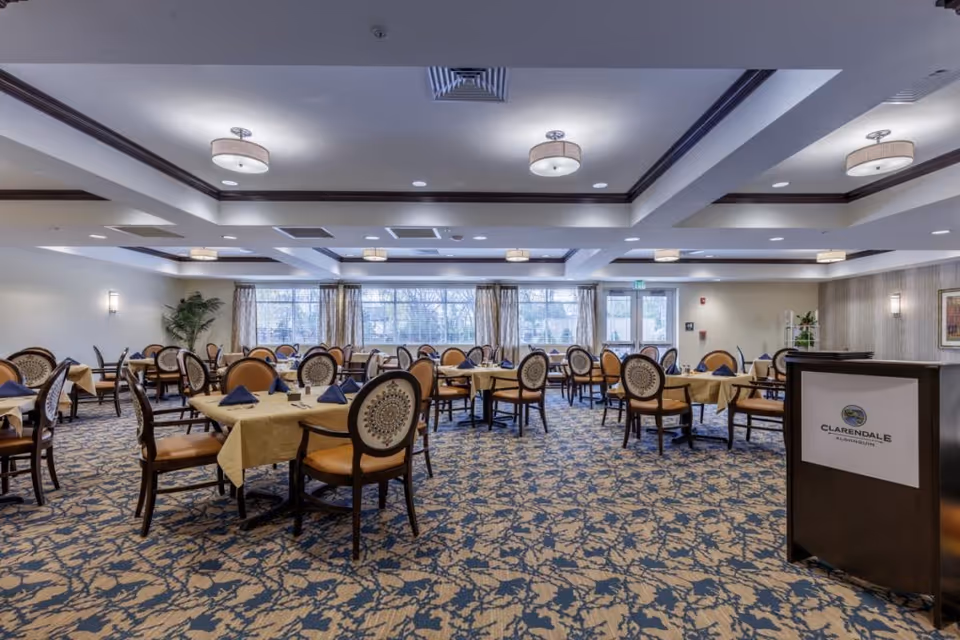 Spacious dining room with multiple tables covered in yellow tablecloths and set with blue napkins. The room features patterned carpet, large windows with curtains, ceiling lights, and a podium with a Clarendale of Algonquin sign.