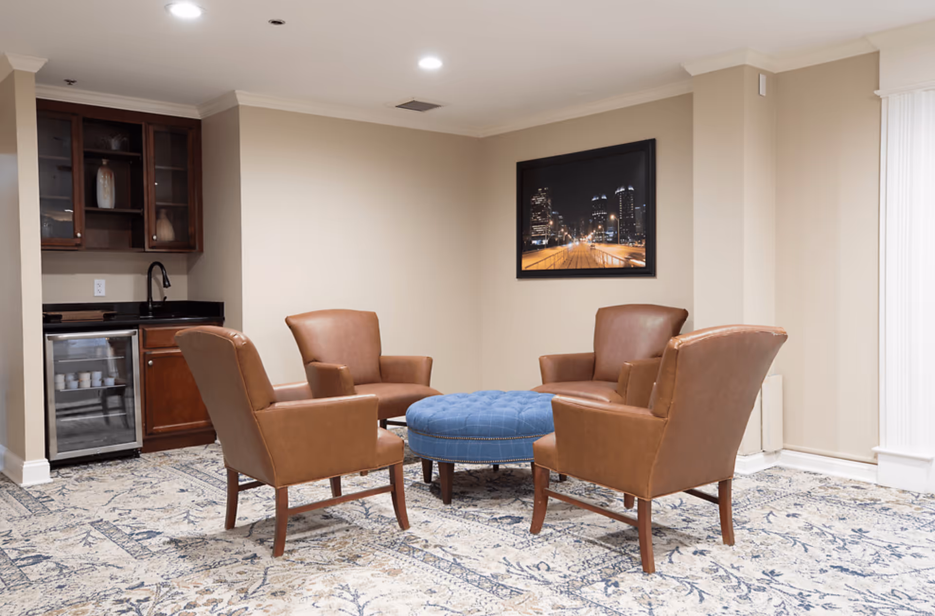 A cozy sitting area with four brown leather armchairs arranged around a round blue upholstered ottoman. The room has beige walls, a patterned carpet, and a small kitchenette with a mini fridge and sink in the corner. A framed cityscape photo hangs on the wall.