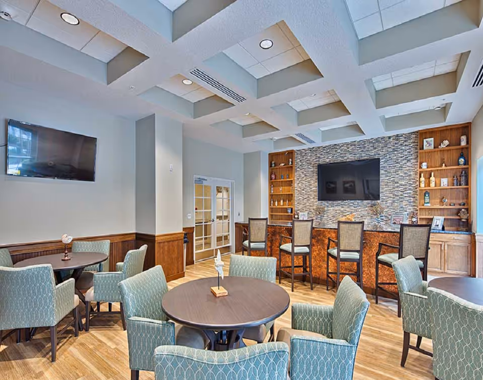 A well-lit common area with round tables surrounded by upholstered chairs, a bar counter with high chairs, built-in wooden shelves with decorative items, and two wall-mounted flat-screen TVs. The ceiling has a modern coffered design with recessed lighting.