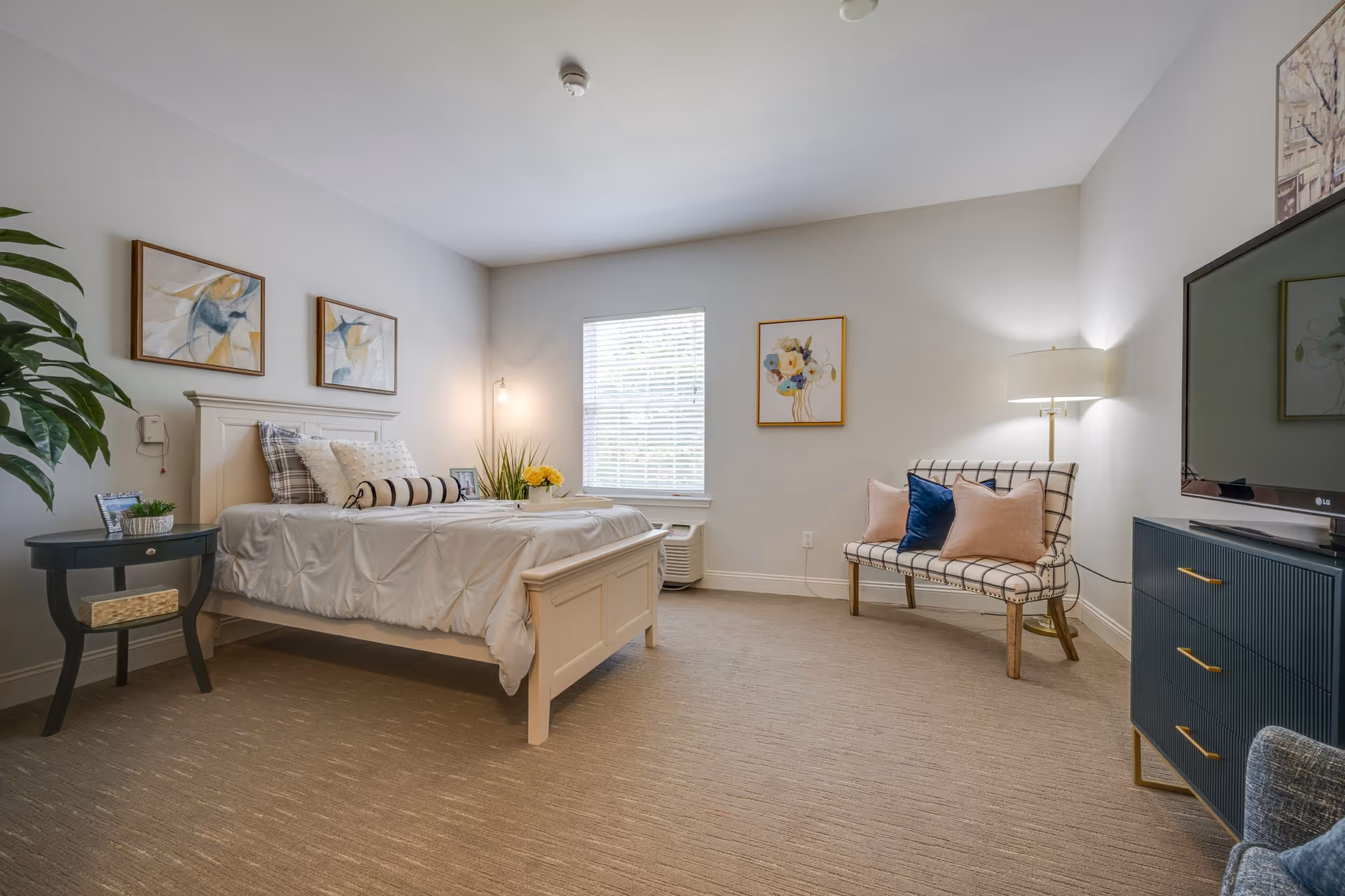 A bright and neatly arranged bedroom in an assisted living facility featuring a white bed with pillows and a bolster, a small black side table with a plant and picture frame, a window with blinds, a patterned loveseat with cushions, a floor lamp, a blue dresser with a flat-screen TV on top, and framed artwork on the walls.